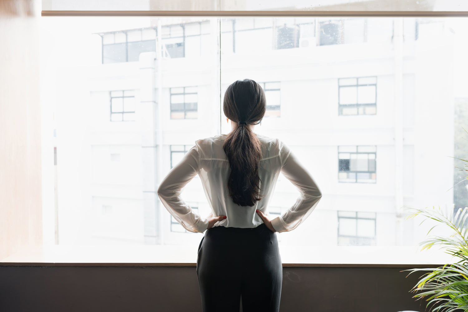 young woman in white shirt with a black skirt on, her hair is in a low ponytail. She is looking out of large window with her hands on hips, appears to be thinking.